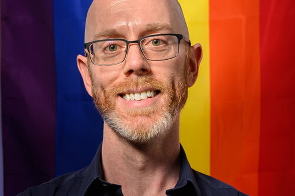 A photo of Dr. Stefan Vogler. He is standing in front of a Progress Pride flag and smiling at the camera.